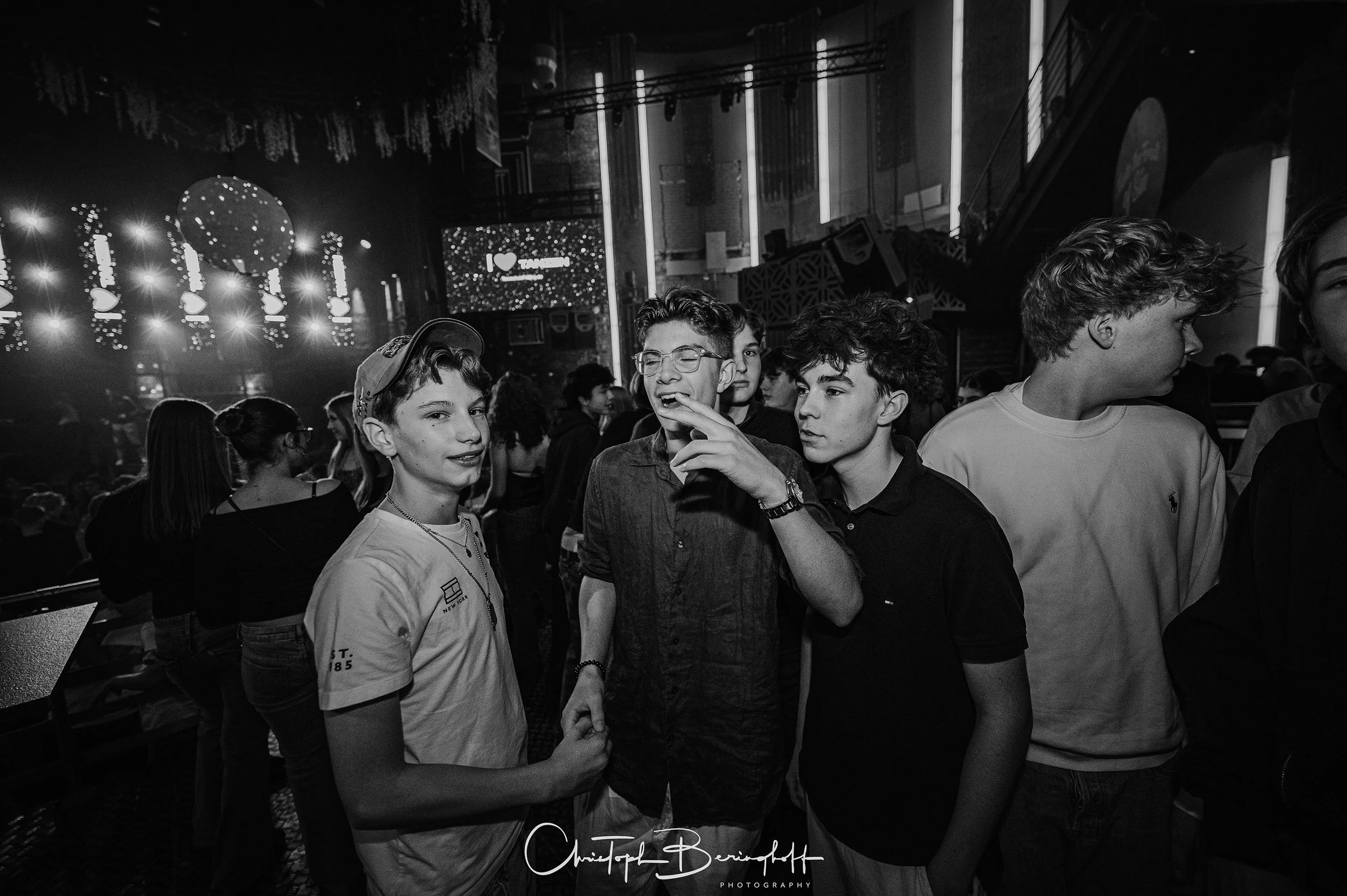 Junge Jugendliche in einer dunklen, festlichen Umgebung, einige lachen und unterhalten sich, mit Lichteffekten im Hintergrund.