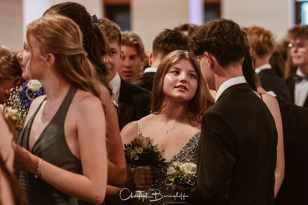 Junge Menschen in formeller Kleidung bei einer Gala oder Feier, einige im Gespräch, mit Blumen in der Hand.