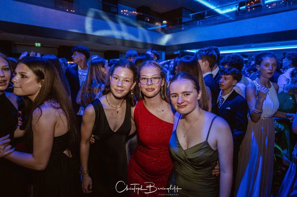 Junge Menschen in formeller Kleidung auf einer Party oder Abschlussfeier, überwiegend Frauen im Vordergrund, Hintergrund mit mehr Menschen und blauer Beleuchtung.