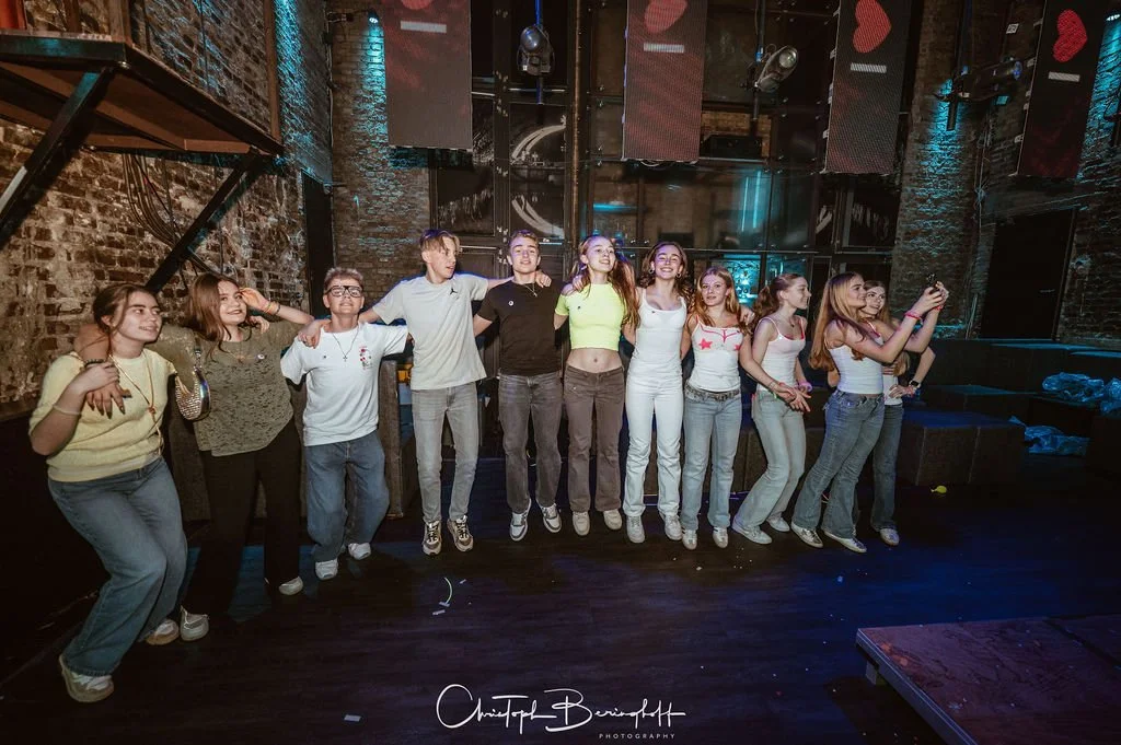 Young girls stand side by side on a stage, some with their arms around each other’s shoulders, posing for photos in a dark, industrial-style setting with brick walls, lighting effects, and jukeboxes in the background.