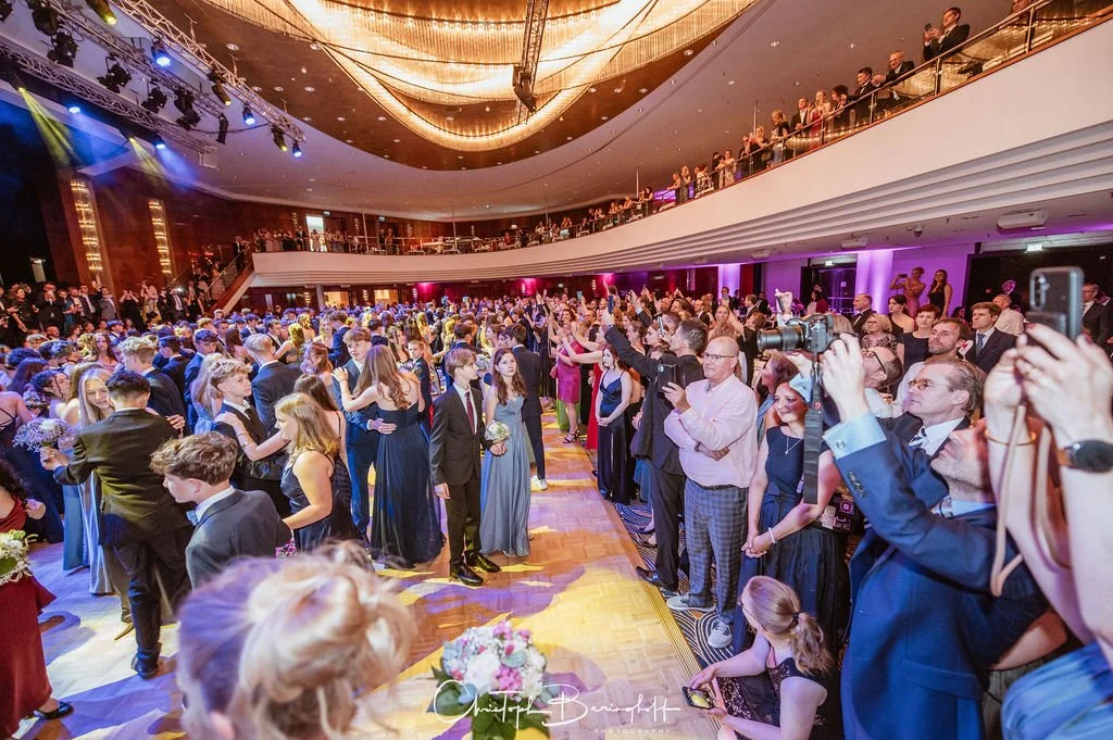 Elegante Gesellschaft bei einer festlichen Veranstaltung im Ballsaal, viele Menschen auf dem Tanzparkett und auf der oberen Galerie, festliche Kleidung, dekorierte Umgebung mit Blumen und einer großen Beleuchtung.