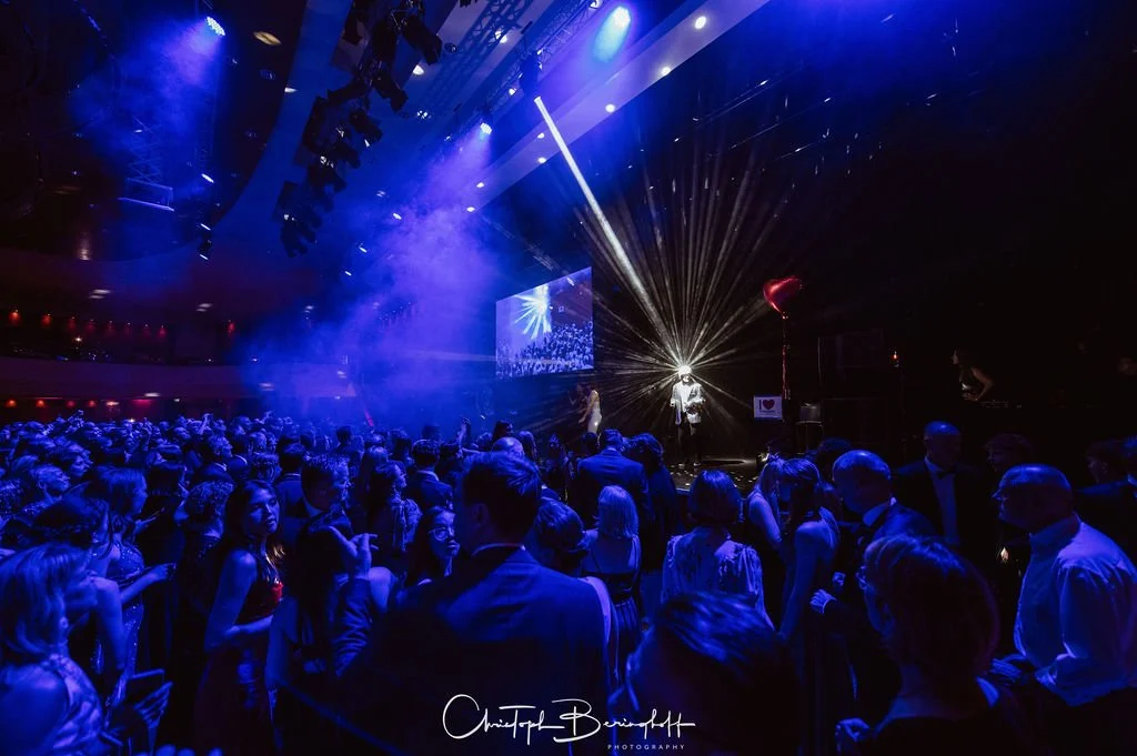 Menschen feiern in einem dunklen Ballsaal mit blauen Lichtern, auf der Bühne sind Show- oder Konzertperformers mit Lichtstrahlen, große Zuschauermenge, viele sehen sich das Geschehen an.