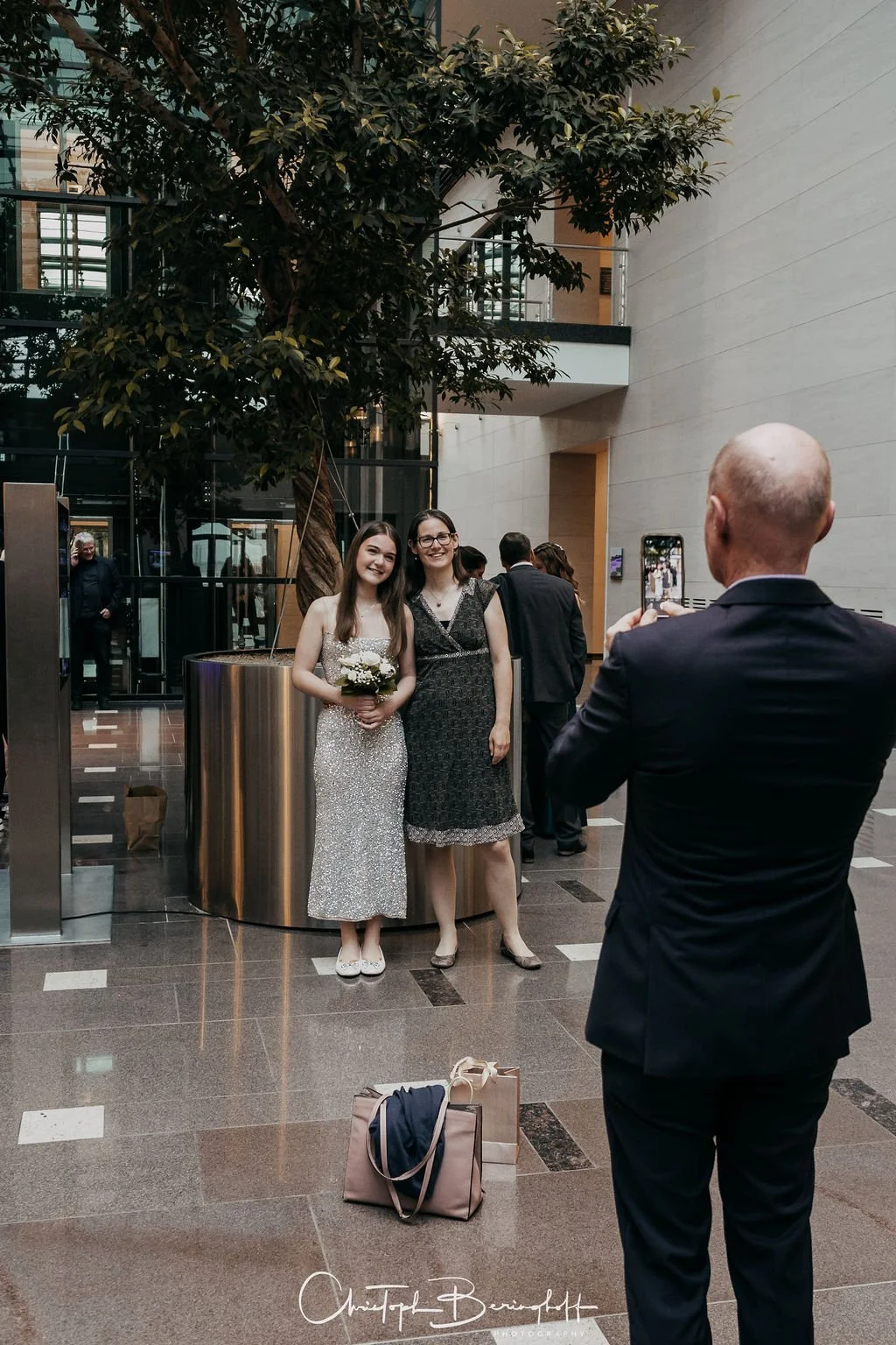 Zwei Frauen, eine in einem funkelnden silbernen Kleid mit Blumenstrauß, posieren für einen Mann, der Fotos mit einem Smartphone macht, in einem modernen Innenraum mit großen Pflanzen und mehreren Menschen im Hintergrund.