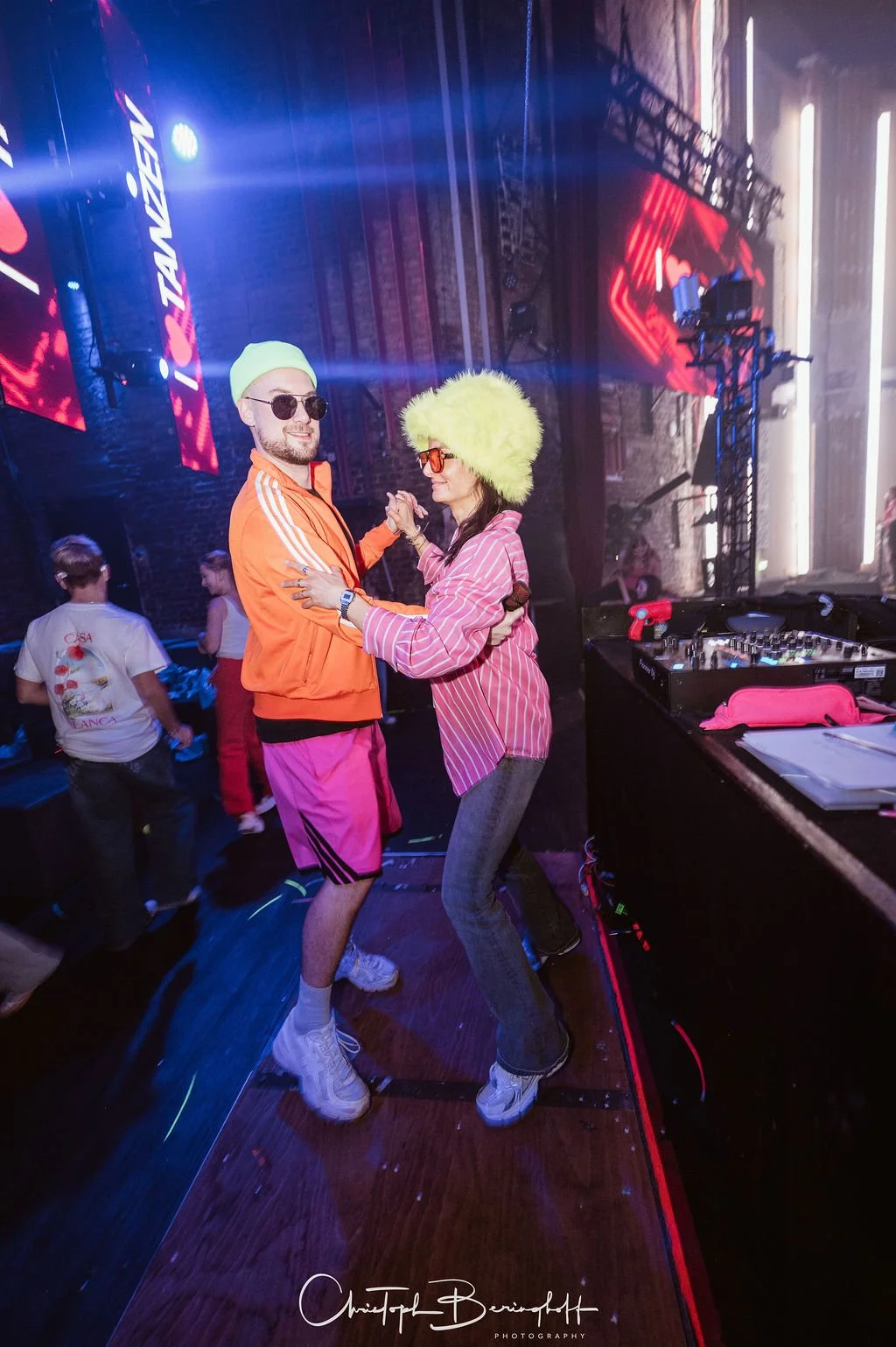 Two people are dancing in a club; both are wearing eye-catching, colorful clothes and glasses. The man is wearing a light green cap, an orange jacket with white stripes on the sleeves, pink shorts, and white sneakers. The woman is wearing a large, yellow F