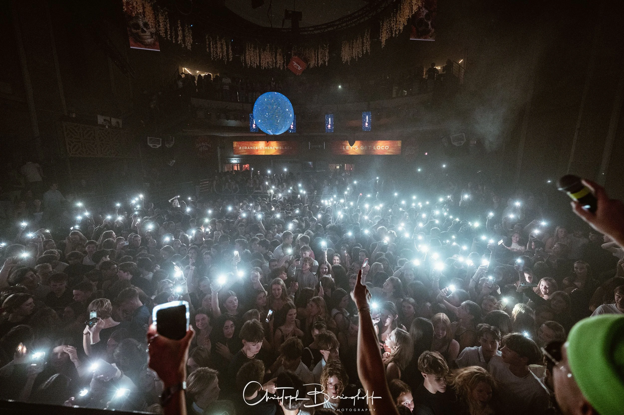 Menschenmenge auf einer Party oder einem Konzert, viele halten ihre Handys mit Taschenlampen hoch, im Hintergrund eine große leuchtende Kugel und ein Bildschirm mit Texten.