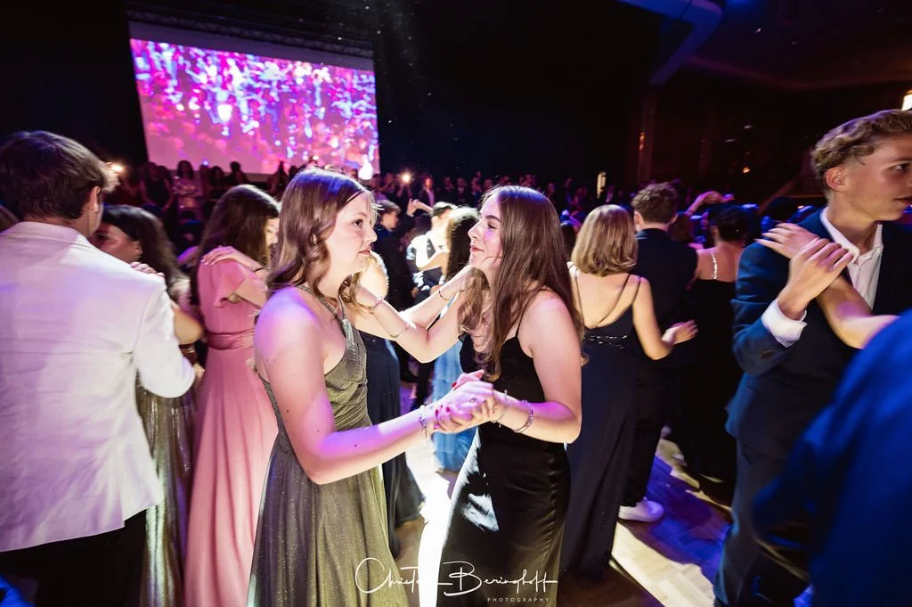 Junge Menschen tanzen auf einer Party oder einem Ball, einige halten sich an den Händen, im Hintergrund eine große leuchtende Leinwand.