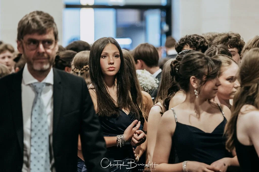 Menschen auf einer formellen Veranstaltung, viele tragen elegante Kleidung, darunter Frauen in schwarzen Kleidern und ein Mann in Anzug mit Krawatte.