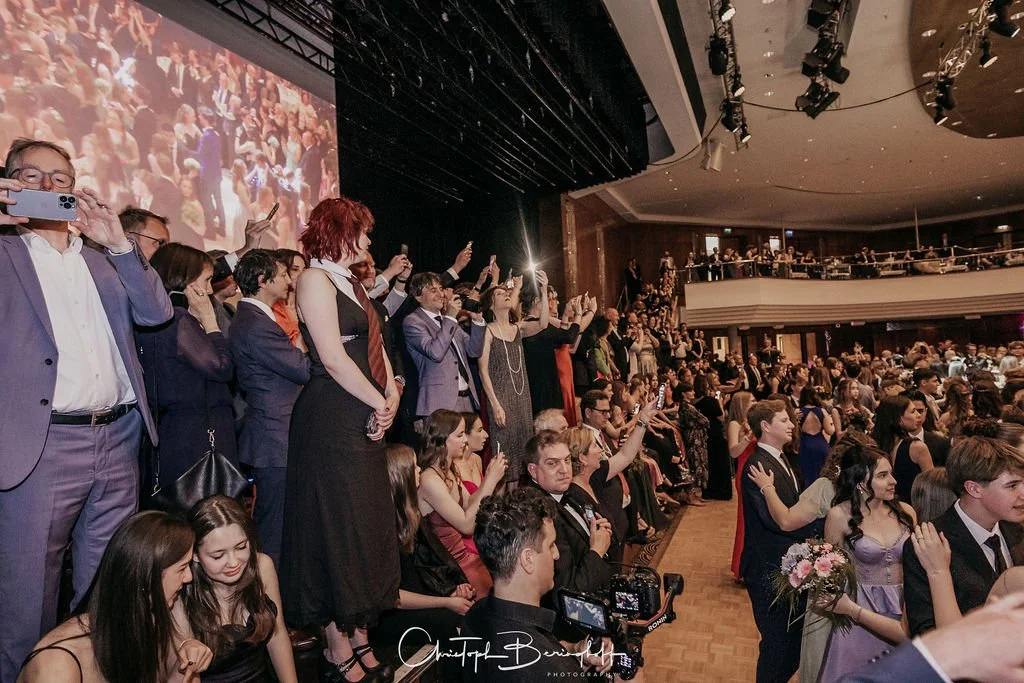 Ein Saal mit einem großen Publikum bei einer Veranstaltung, viele Menschen stehen und sitzen, einige sind am Fotografieren oder Filmen, eine große Leinwand im Hintergrund zeigt ein Bild der Menge.