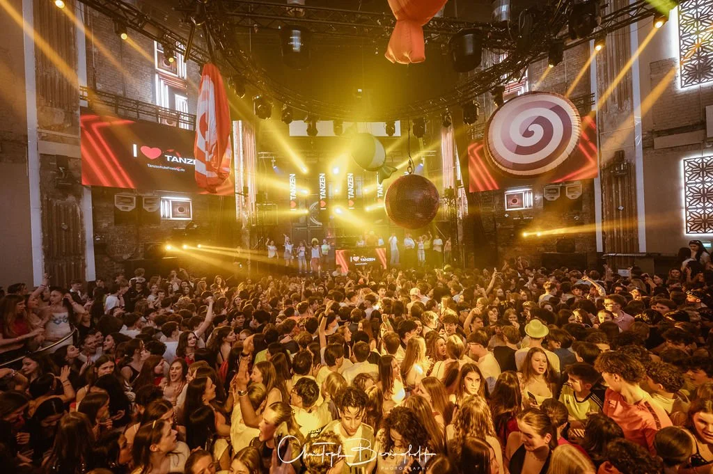 A large crowd at a party or concert in a venue with a stage and colorful lighting effects.