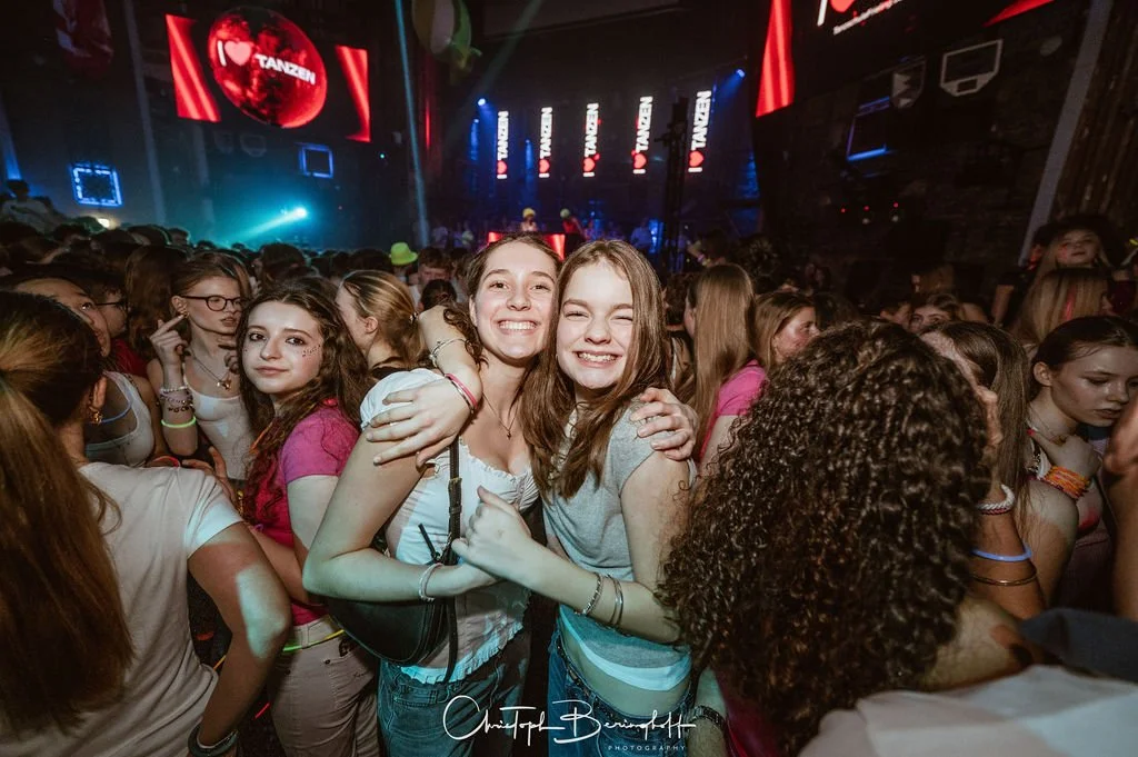Two smiling young women are hugging each other at a club party, surrounded by a crowd of young people, with a large stage set in the background.