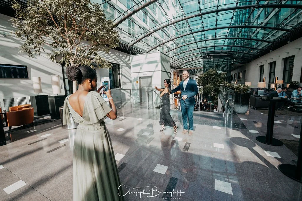 Ein Mann und eine Frau tanzen in einem modernen, glasüberdachten Gebäude, während eine andere Frau ein Foto von ihnen macht, im Hintergrund sitzen Menschen und es gibt Pflanzen.
