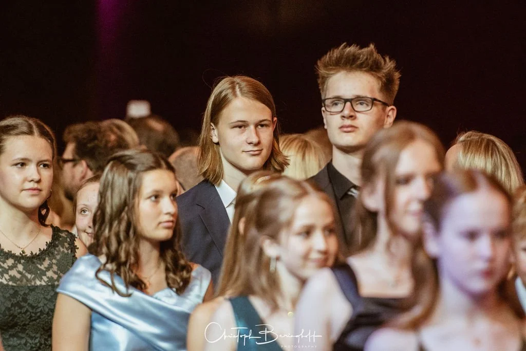 Junge Menschen auf einer Feier, die meisten tragen formelle Kleidung. Zwei Personen, ein Junge mit Brille und ein Mädchen, stehen im Hintergrund und schauen nach vorne.