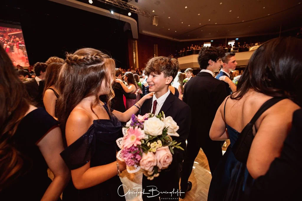 Menschen tanzen bei einer formellen Veranstaltung, Frauen in eleganten Kleidern, Männer in Anzügen, einem Jungen und einem Mädchen, das ein Blumenbouquet hält.