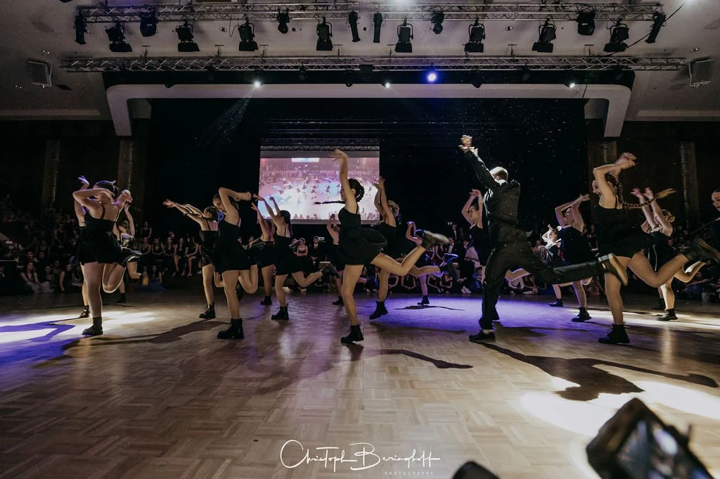 Tanzgruppe aus jungen Frauen und Männern performing auf der Bühne, in schwarzen Outfits, im Mittelpunkt, mit Publikum im Hintergrund, dunkler Raum, Bühnenlicht, Holzparkett