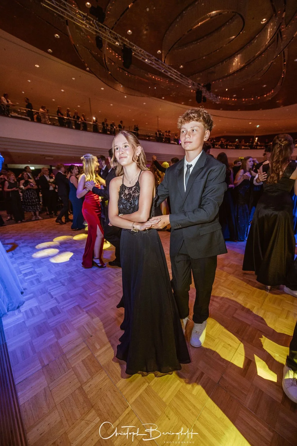 Junge Paare tanzen auf einer festlichen Tanzfläche bei einer formellen Veranstaltung, viele Gäste in eleganter Kleidung, dekorierte Räume mit buntem Licht.