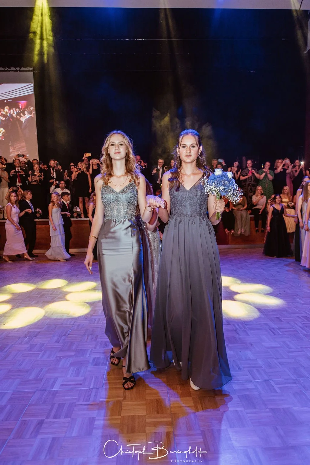Zwei Frauen in eleganten Abendkleidern tanzen bei einer Feier auf der Tanzfläche, umgeben von weiteren Gästen im Hintergrund.