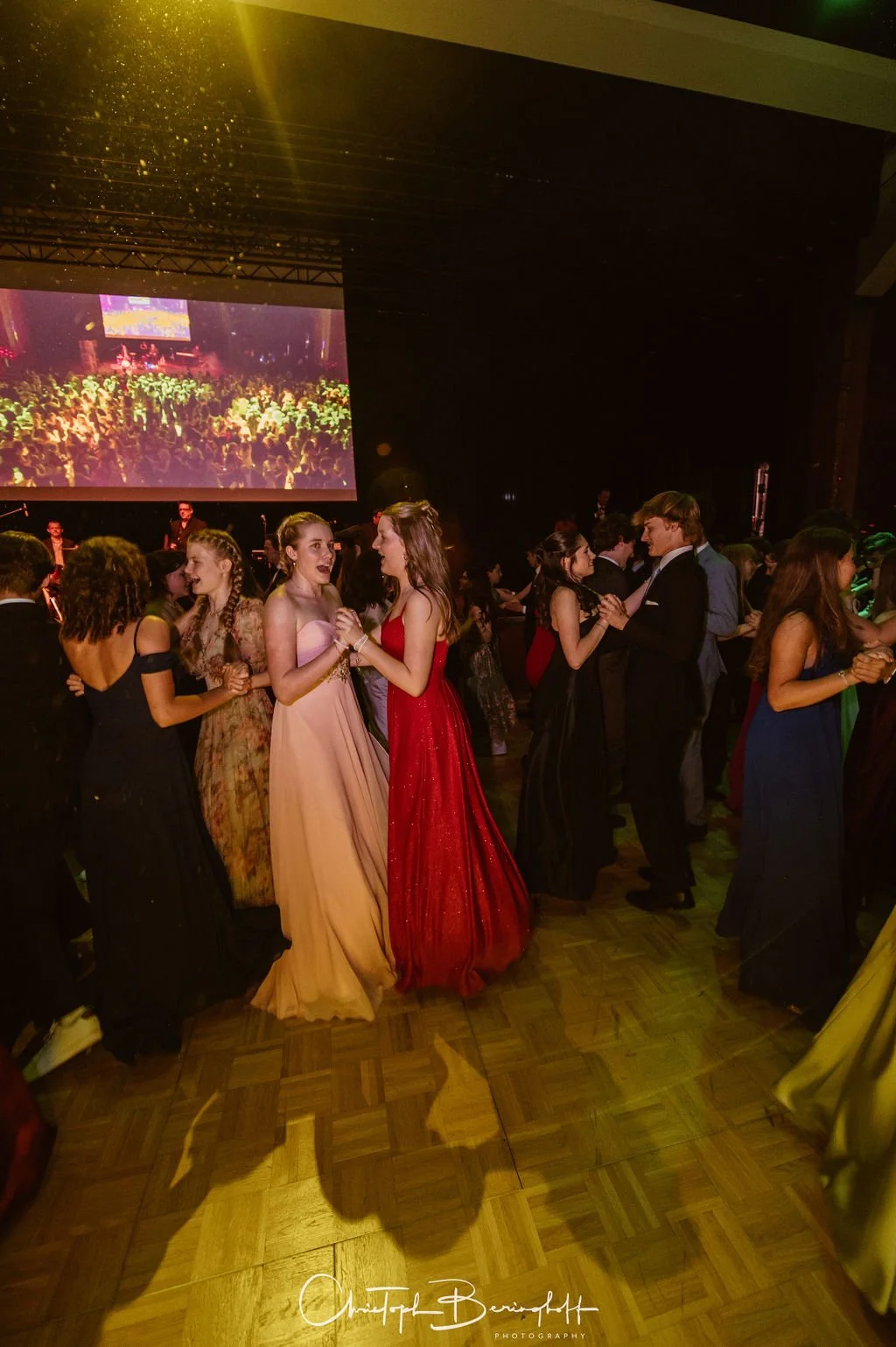 Junge Menschen tanzen bei einer Gala oder Ball, im Hintergrund eine Bühne mit großem Bildschirm. Elegante Kleidung, festliche Atmosphäre.