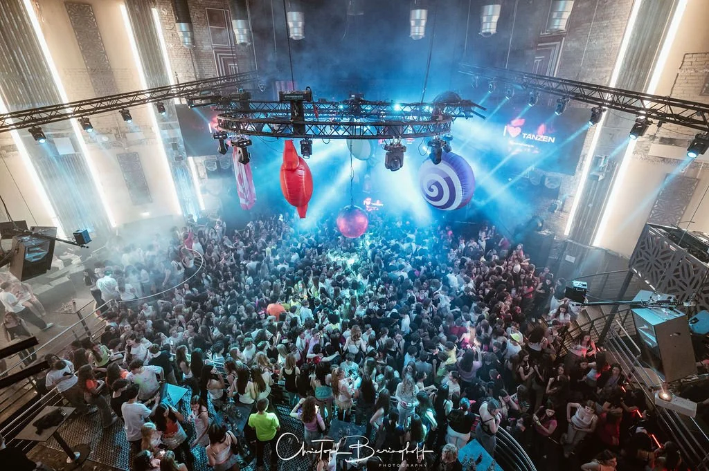 A large club with lots of people dancing, colorful and bright light shows, large decorations hanging from the ceiling, and dance performances in the background.