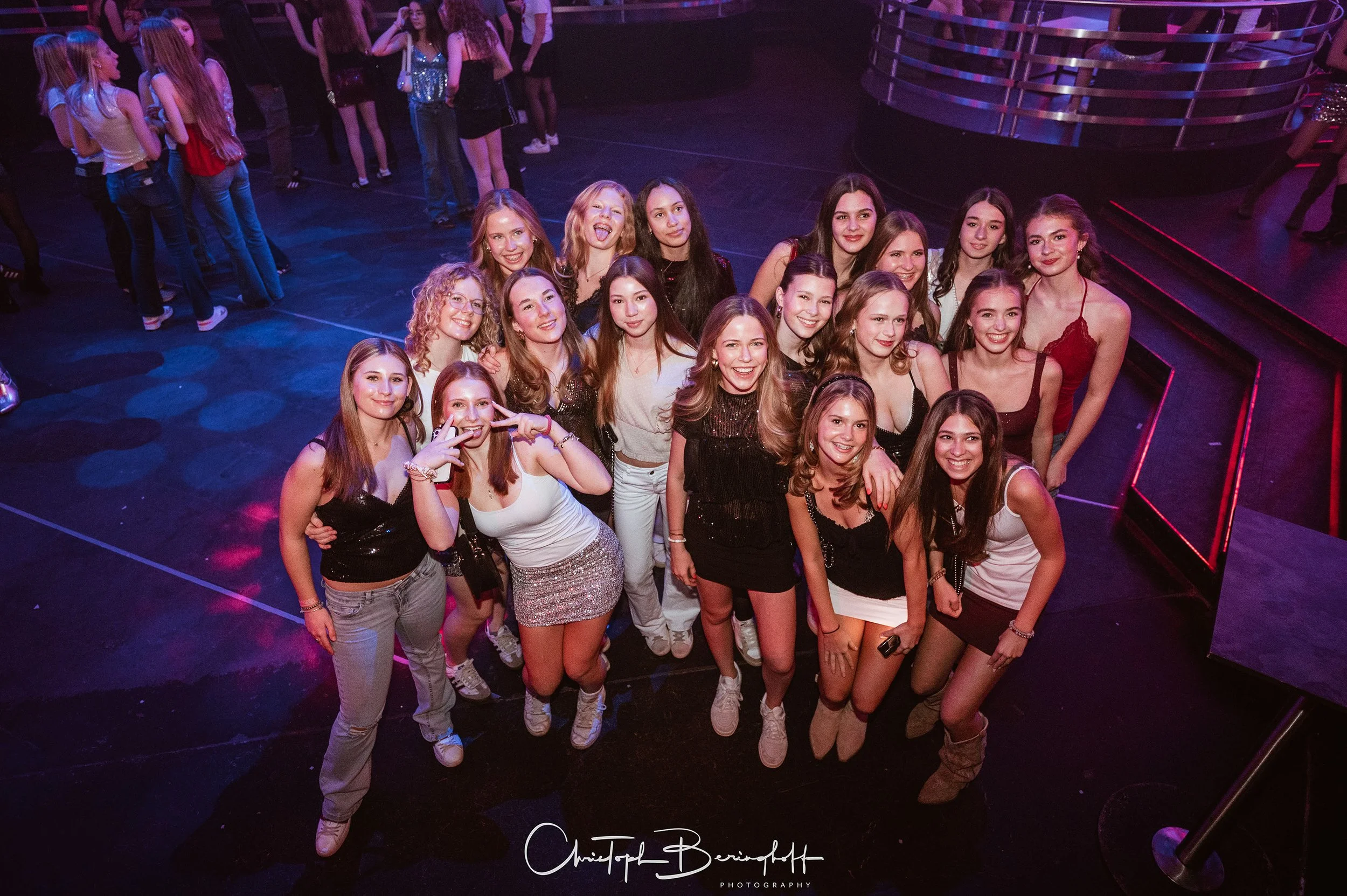 Eine Gruppe junger Frauen, die auf einer Party lachen und posieren, vor einem dunklen Hintergrund mit bunten Lichtern.