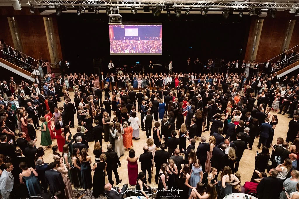 Großgruppe von Leuten bei einer formellen Veranstaltung, die auf einer Tanzfläche tanzen, im Hintergrund ein Bildschirm und eine Band.