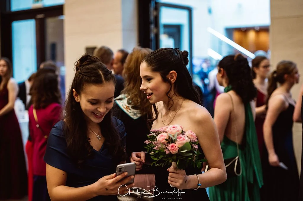 Zwei Frauen schauen jubelnd auf ein Smartphone, eine hält einen Blumenstrauß bei einer festlichen Veranstaltung.