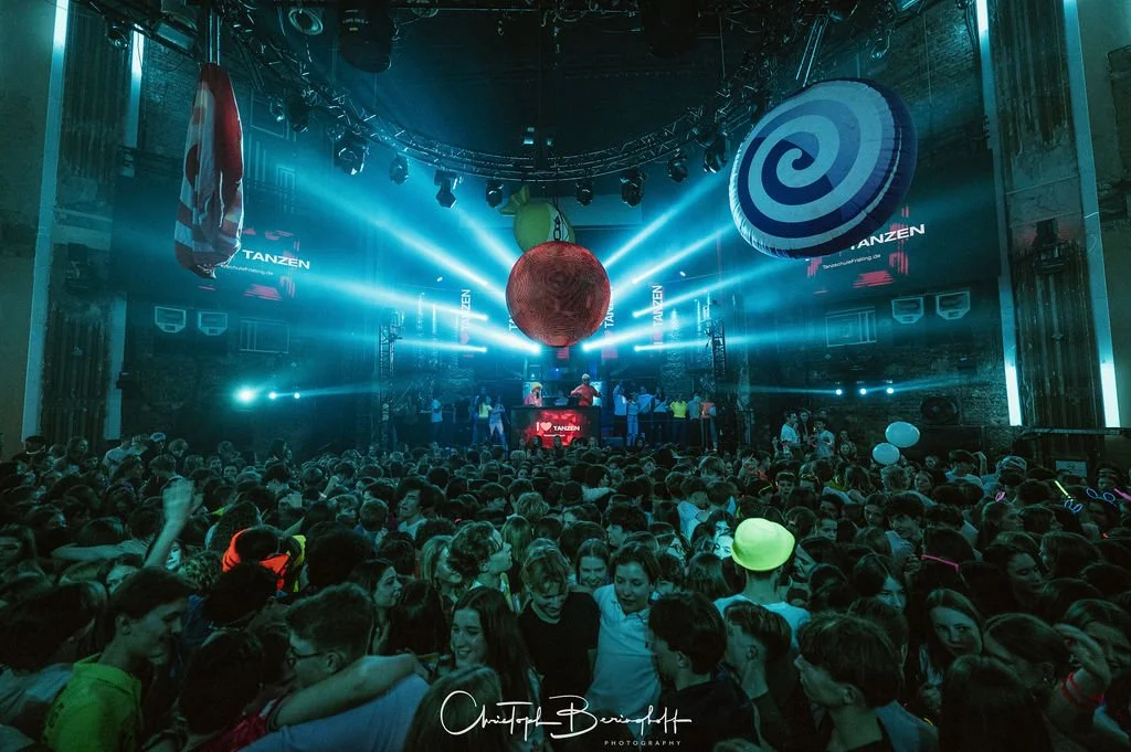 A crowd at a party with colorful LED lights, large inflatable decorations, and a DJ booth on the stage in the background.