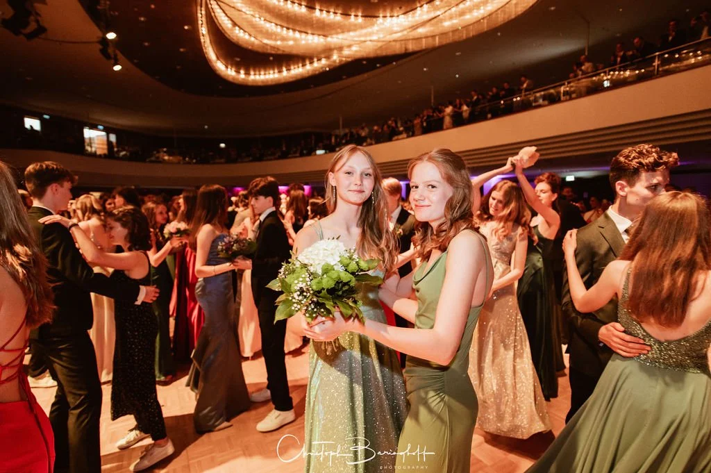 Menschen tanzen auf einer eleganten Party im Ballsaal, zwei Frauen in der Mitte halten einen Blumenstrauß.