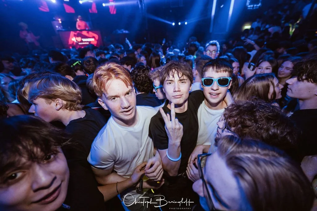 A group of young people at a party or concert in a dark room with colorful lights, most of them in groups and smiling.