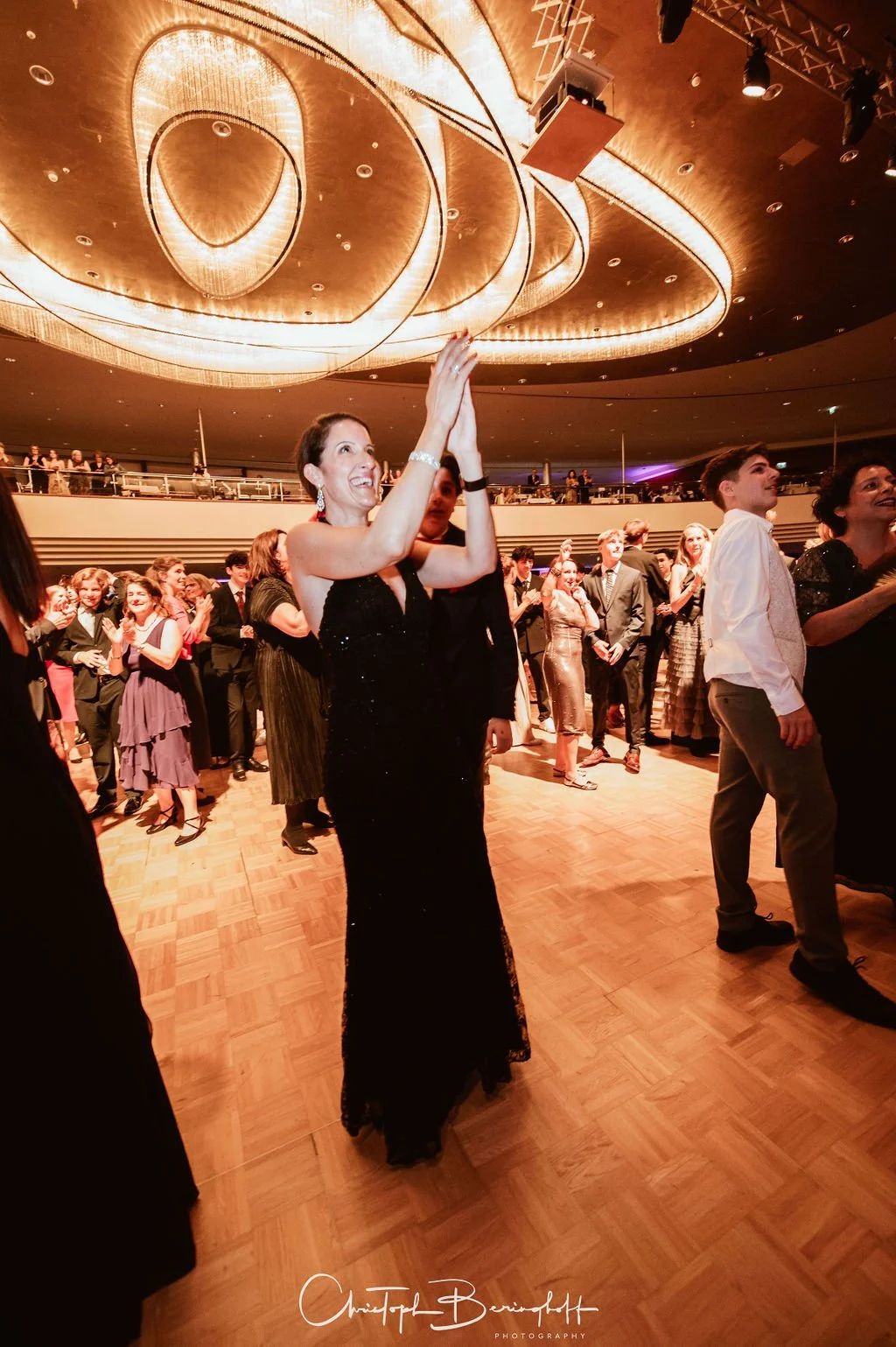Ein Model in einem schwarzen, eleganten Abendkleid applaudiert auf einer festlichen Veranstaltung, umgeben von anderen Gästen im Anzug und Abendkleid, in einem festlich beleuchteten Saal mit großem, moderner Kronleuchter an der Decke.