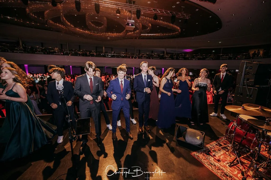 Junge Erwachsene in eleganten Kleidern und Anzügen tanzen auf einer Tanzfläche bei einer Party in einem großen Veranstaltungssaal mit einem Golddekor in der Decke.