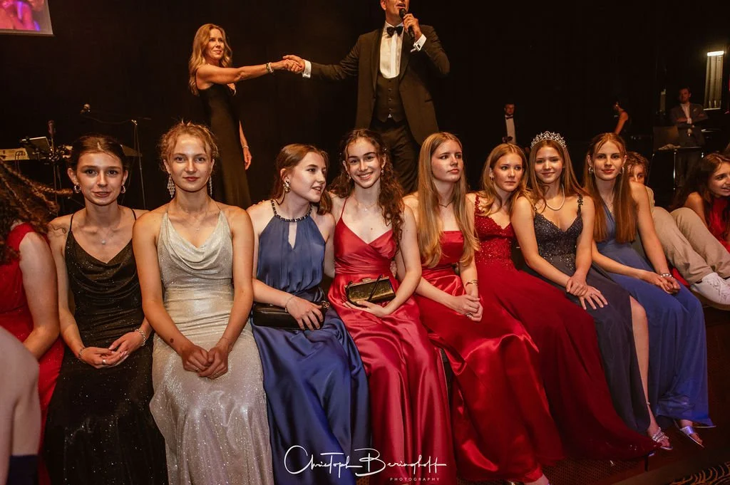 Gruppe von jungen Frauen in eleganten Abendkleidern bei einer Preisverleihung, im Hintergrund eine Bühne mit einem Mann, der einen Preis übergibt, und ein Pianist in einer Ecke.