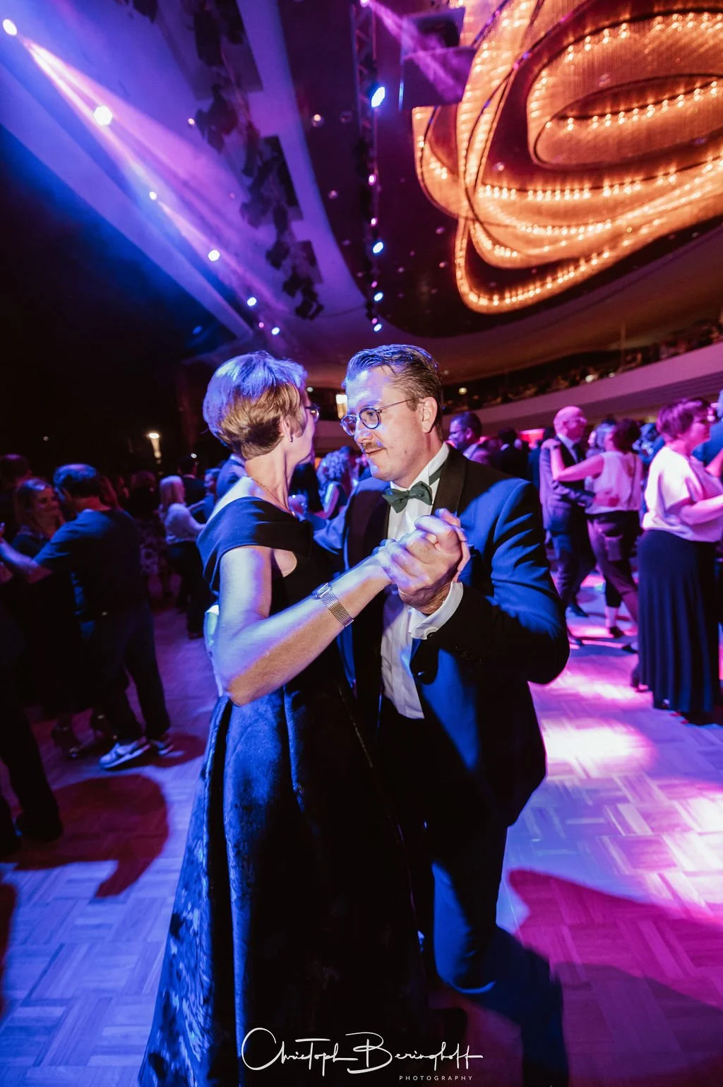Zwei Menschen tanzen in einem elegant beleuchteten Ballsaal. Der Mann trägt einen schwarzen Anzug mit Fliege, die Frau trägt ein langes, dunkles Kleid.