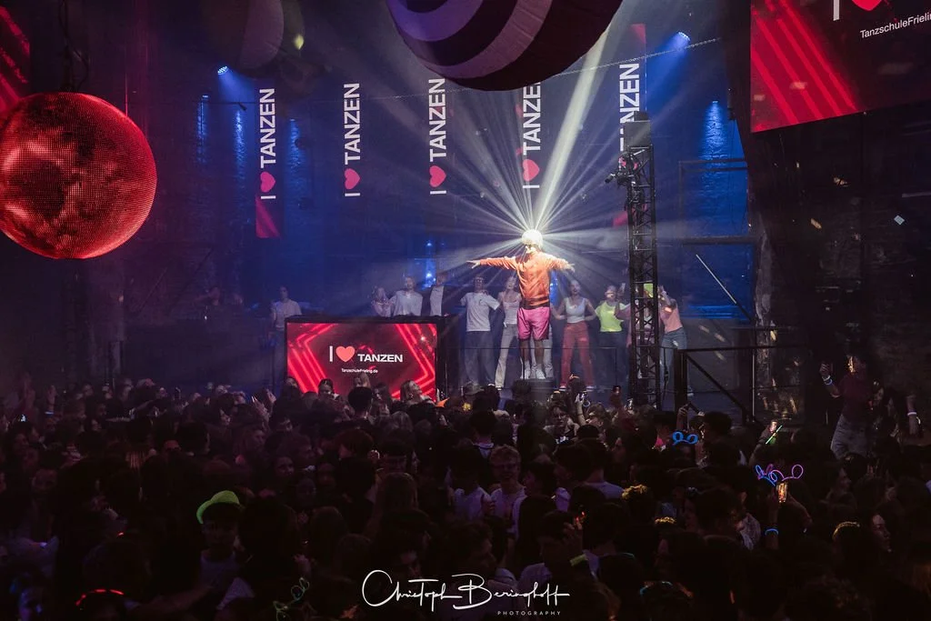 A dance party with people in a dark room, colorful lights, a disco ball, a stage with an energetic show, signs that say "I love dancing," and many guests wearing glowing accessories.
