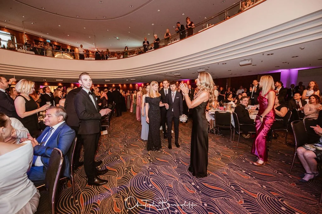 Elegante Gesellschaft bei einer formellen Veranstaltung in einem modern gestalteten Saal, einige Menschen applaudieren, andere sitzen an Tischen, Mädchen in Abendkleidern, junge Männer in Anzügen.