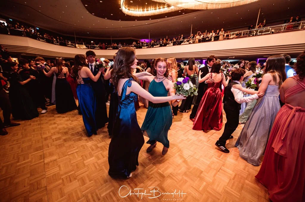 Menschen tanzen auf einer festlichen Veranstaltung in einem großen Saal, einige halten Blumen, mehrere Ebenen mit Zuschauern im Hintergrund.
