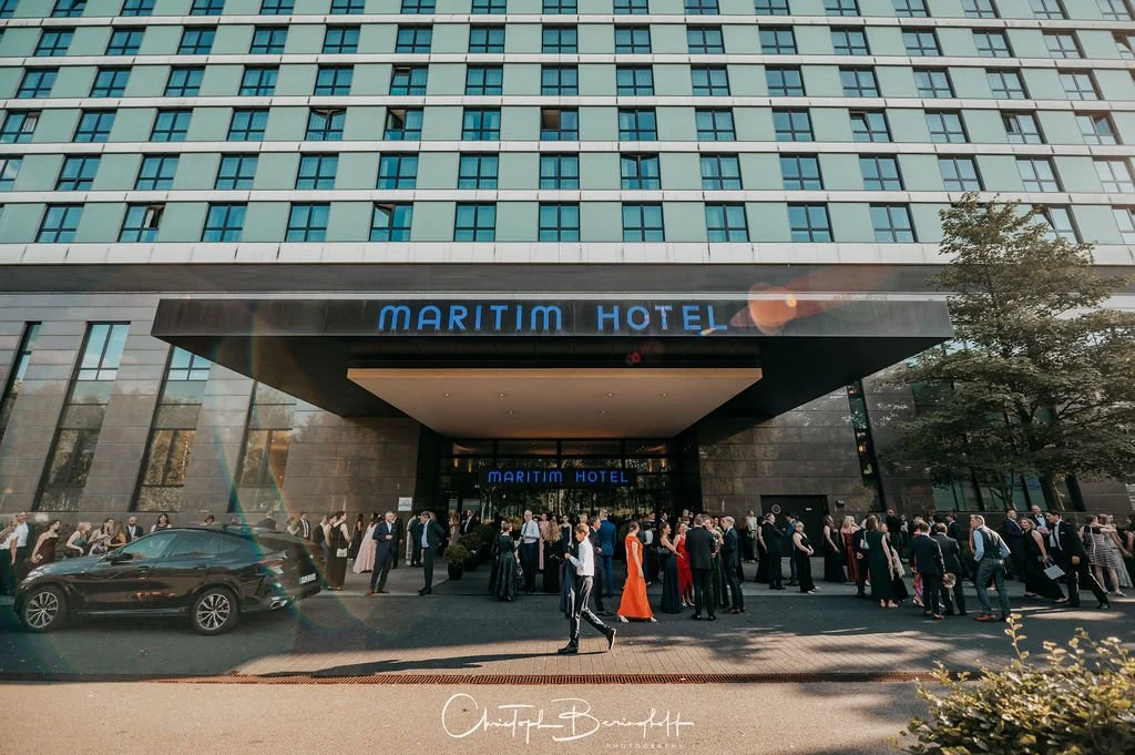 Das Bild zeigt das Maritim Hotel mit einer Stammgäste-Warteschlange vor dem Eingangsbereich, im Vordergrund ein dunkler Auto und Menschen in formeller Kleidung.