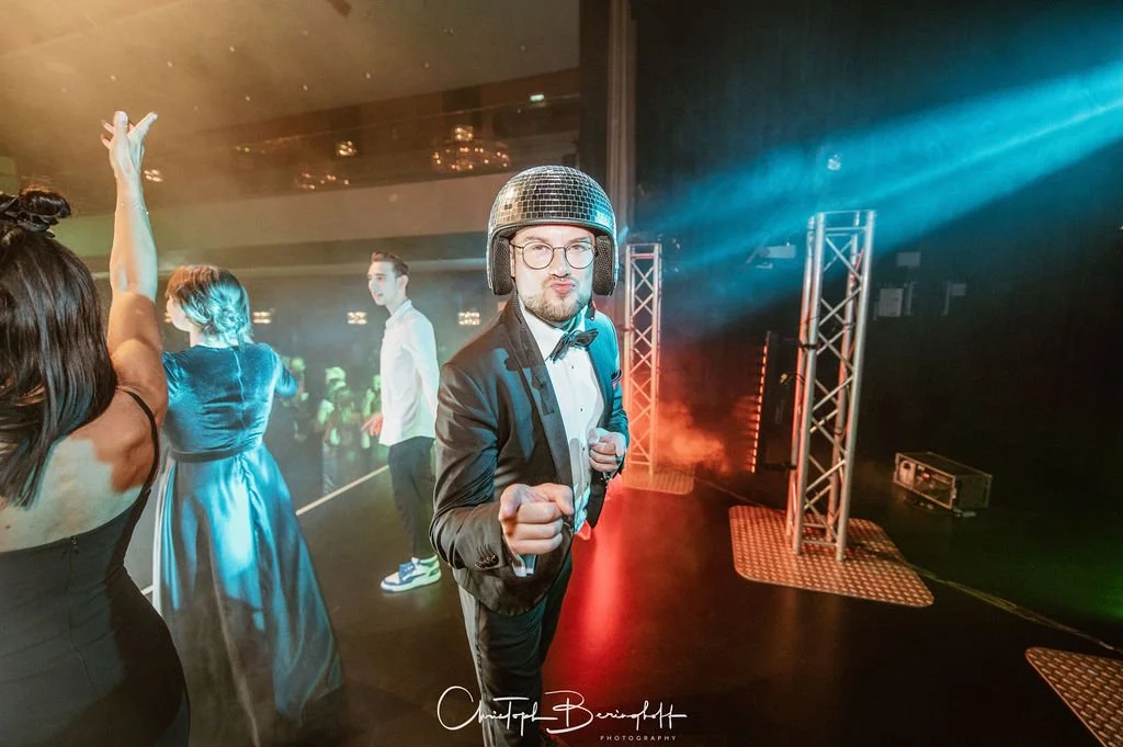 Mann in einem Smoking mit Discokugelhelm posiert auf der Bühne bei einer Party, umgeben von tanzenden Menschen in eleganter Kleidung, im Hintergrund bunte Lichteffekte.