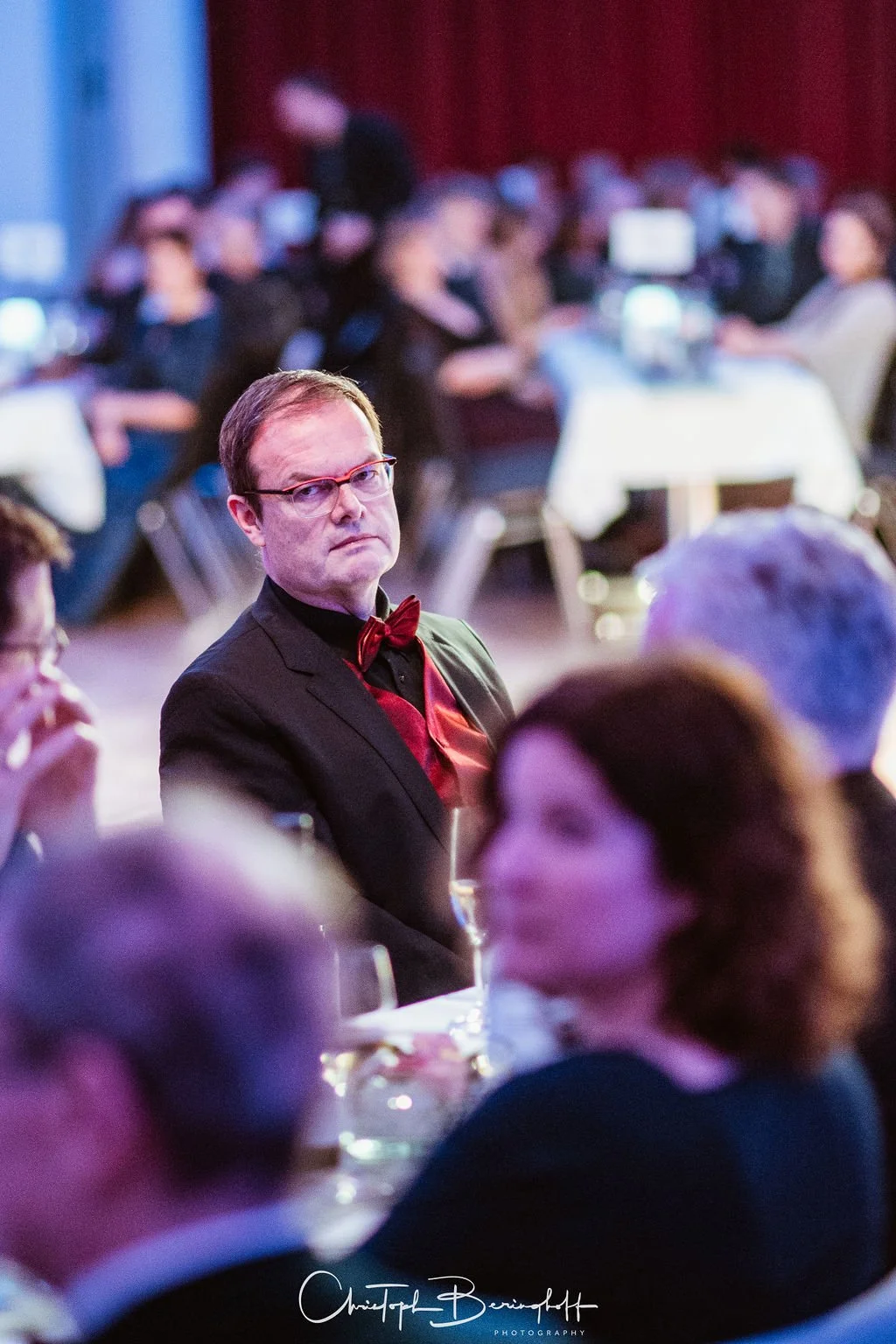 Ein Mann in formellem schwarzen Anzug mit roten Akzenten, roter Fliege und Hemd, sitzt bei einer Veranstaltung in einem Saal, umgeben von anderen Personen, die an Tischen sitzen.