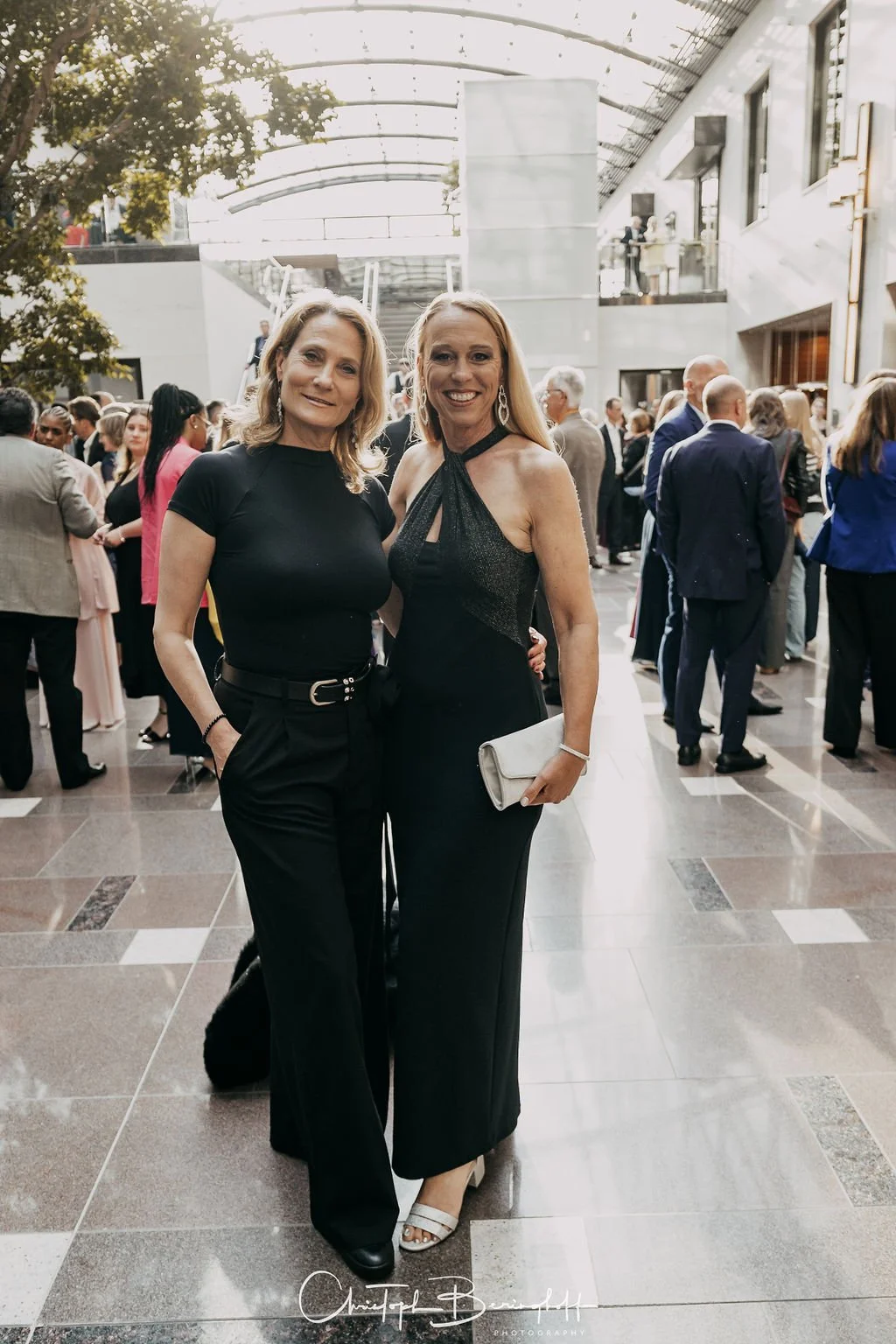 Zwei Frauen in eleganter Kleidung bei einer formellen Veranstaltung, im Hintergrund viele weitere Gäste, im modernen, hellen Innenraum.