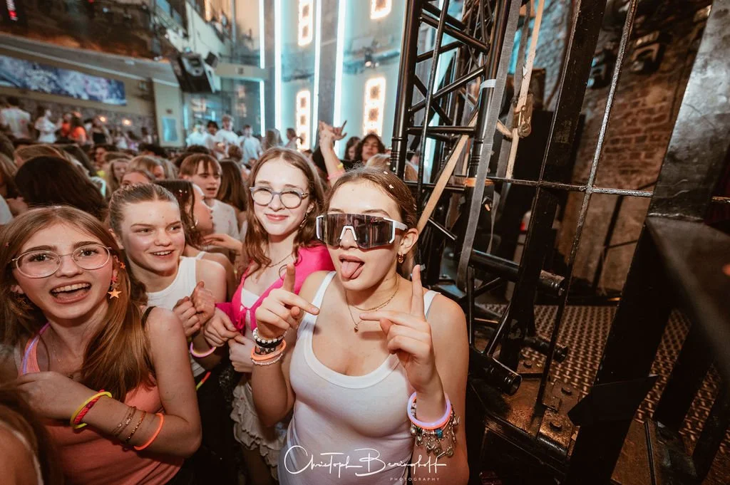 A group of young women are partying at a club; some are wearing colorful sunglasses and jewelry, one girl is striking a pose and making a peace sign, and there are many other partygoers in the background.