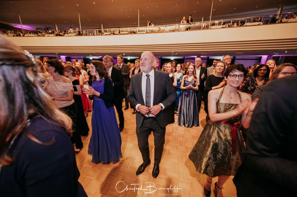Menschen tanzen und feiern auf einer Tanzfläche bei einer formellen Veranstaltung in einem festlich beleuchteten Saal.