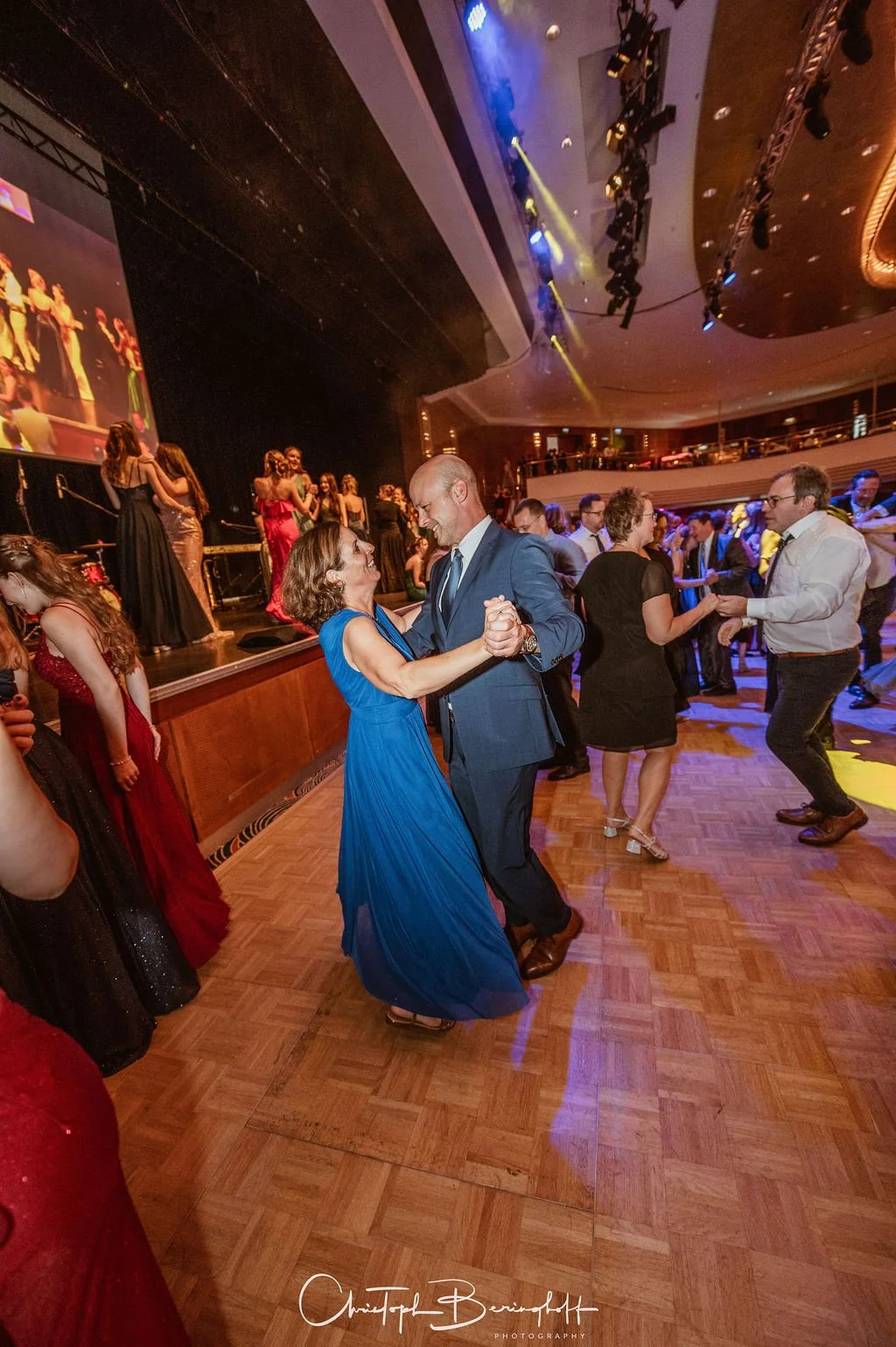 Paare tanzen auf einer festlichen Veranstaltung in einem großen Saal mit einer Tanzfläche, auf einer Bühne laufen junge Ballettänzer. Die Atmosphäre ist lebendig und festlich, mit bunten Lichtern und einer großen Leinwand im Hintergrund.