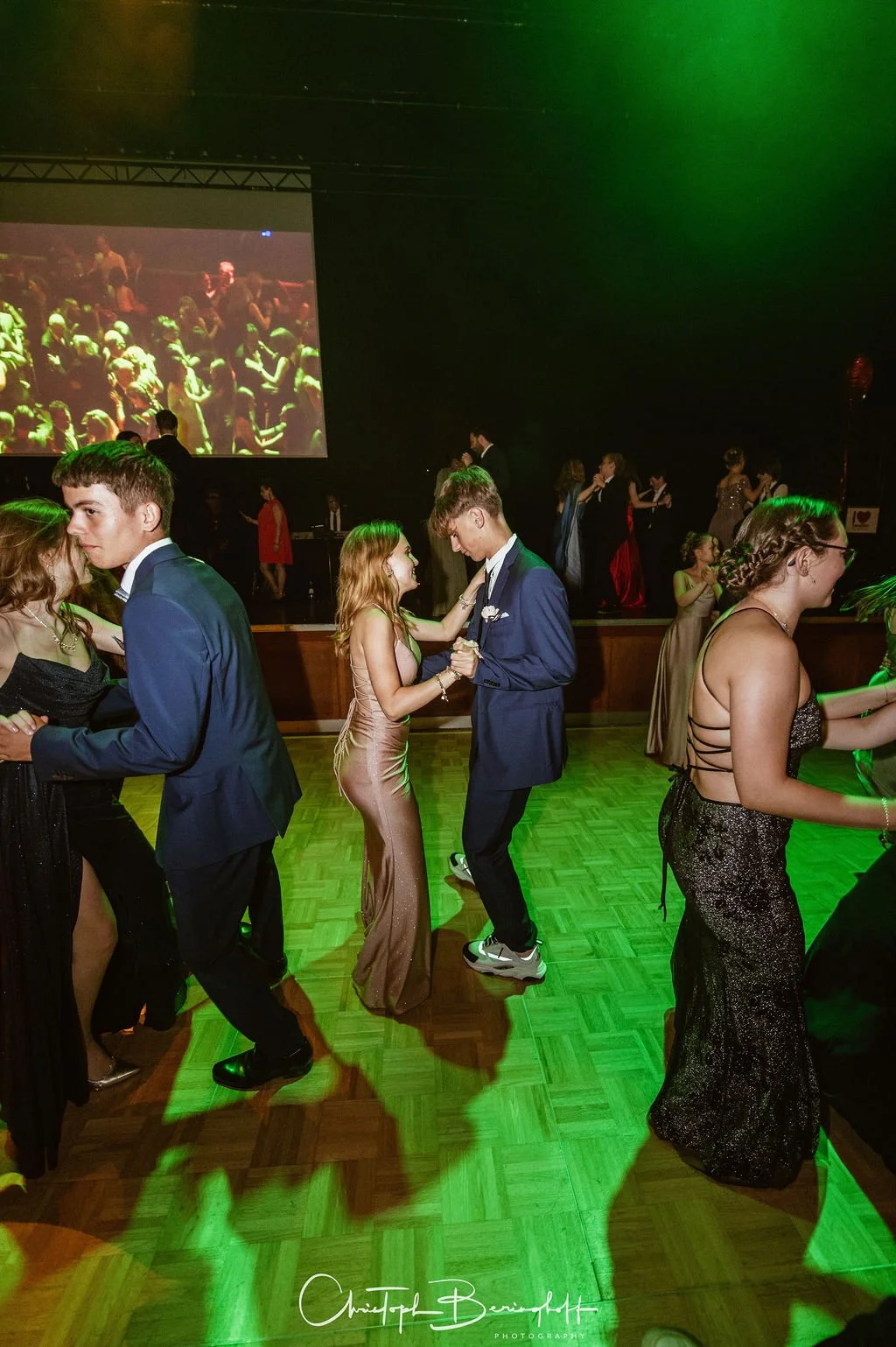 Junge Leute tanzen auf einem festlich dekorierten Tanzball im eleganten Abendkleid und Anzug, mit einem projecting auf einer großen Leinwand im Hintergrund.