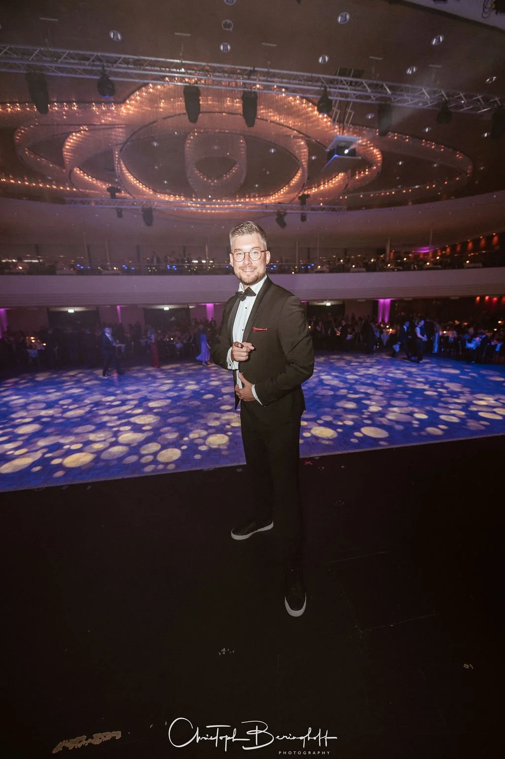 Ein Mann in formeller Kleidung, schwarzer Anzug, Fliege und Brille, steht auf einer Bühne bei einer Gala oder Ball, im Hintergrund sind Tische mit Gästen und eine große Tanzfläche mit buntem Lichtmuster.