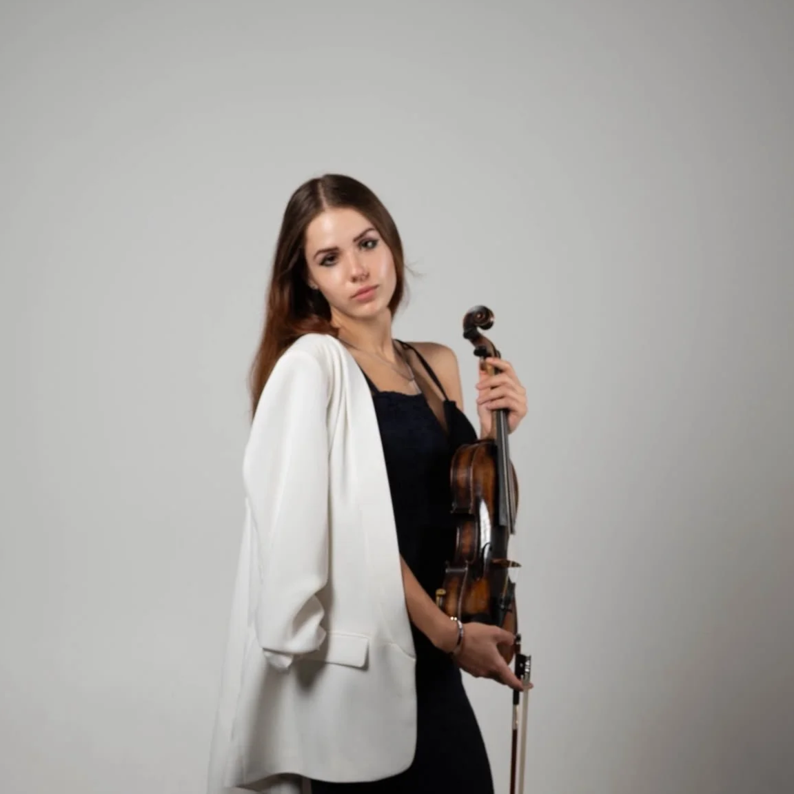 Kateryna with long brown hair holding a violin, dressed in a black outfit with a white blazer and standing against a plain gray background.