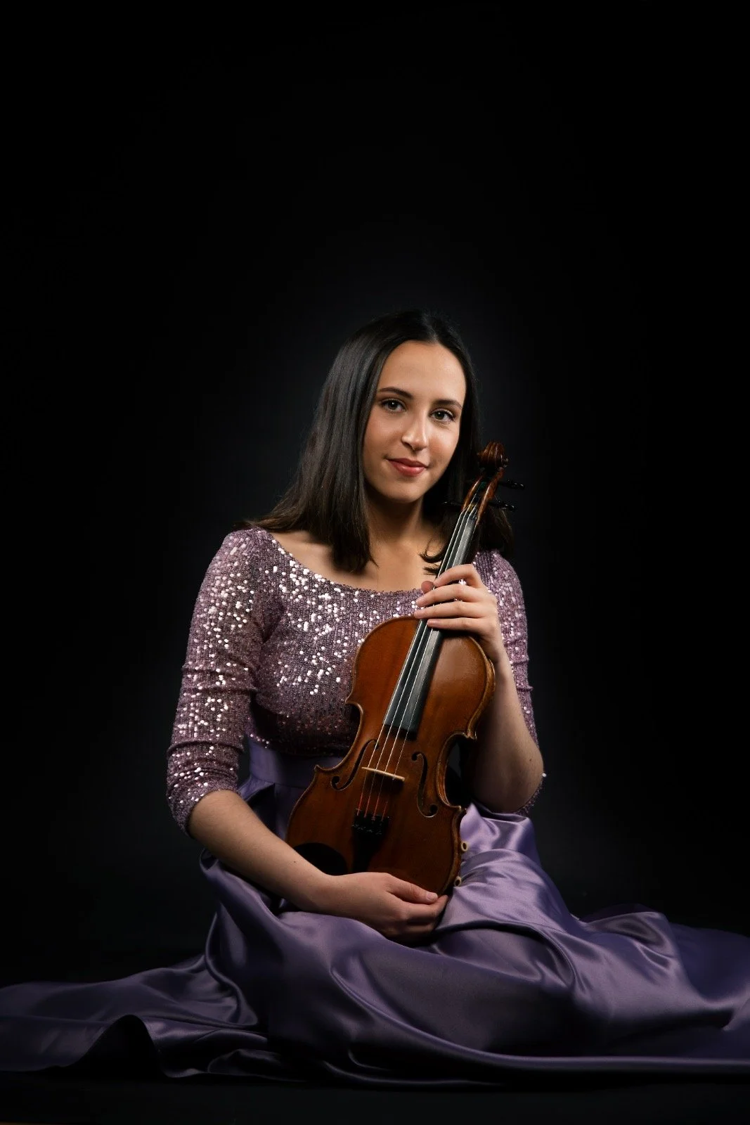 Nina with shoulder-length dark hair, sitting on the floor with a purple satin dress, holding a violin, against a black background.
