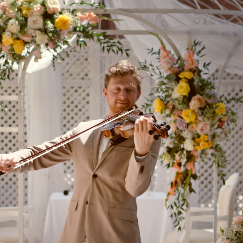 Nikolaus in a tan suit playing a violin at a decorated outdoor wedding ceremony with floral arrangements and a white lattice background.