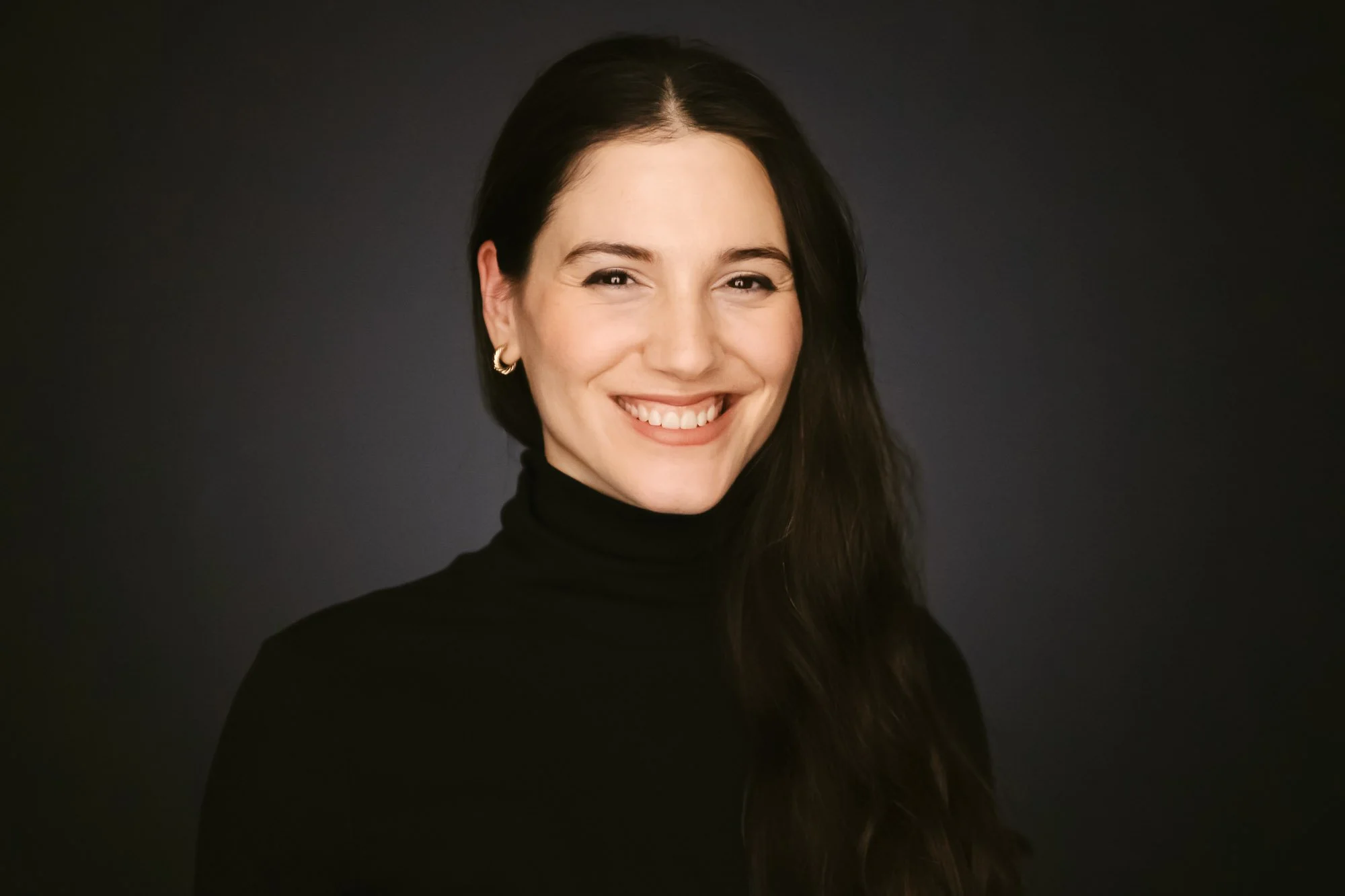 Danae smiling, wearing a black turtleneck and earrings, with dark hair against a dark background.