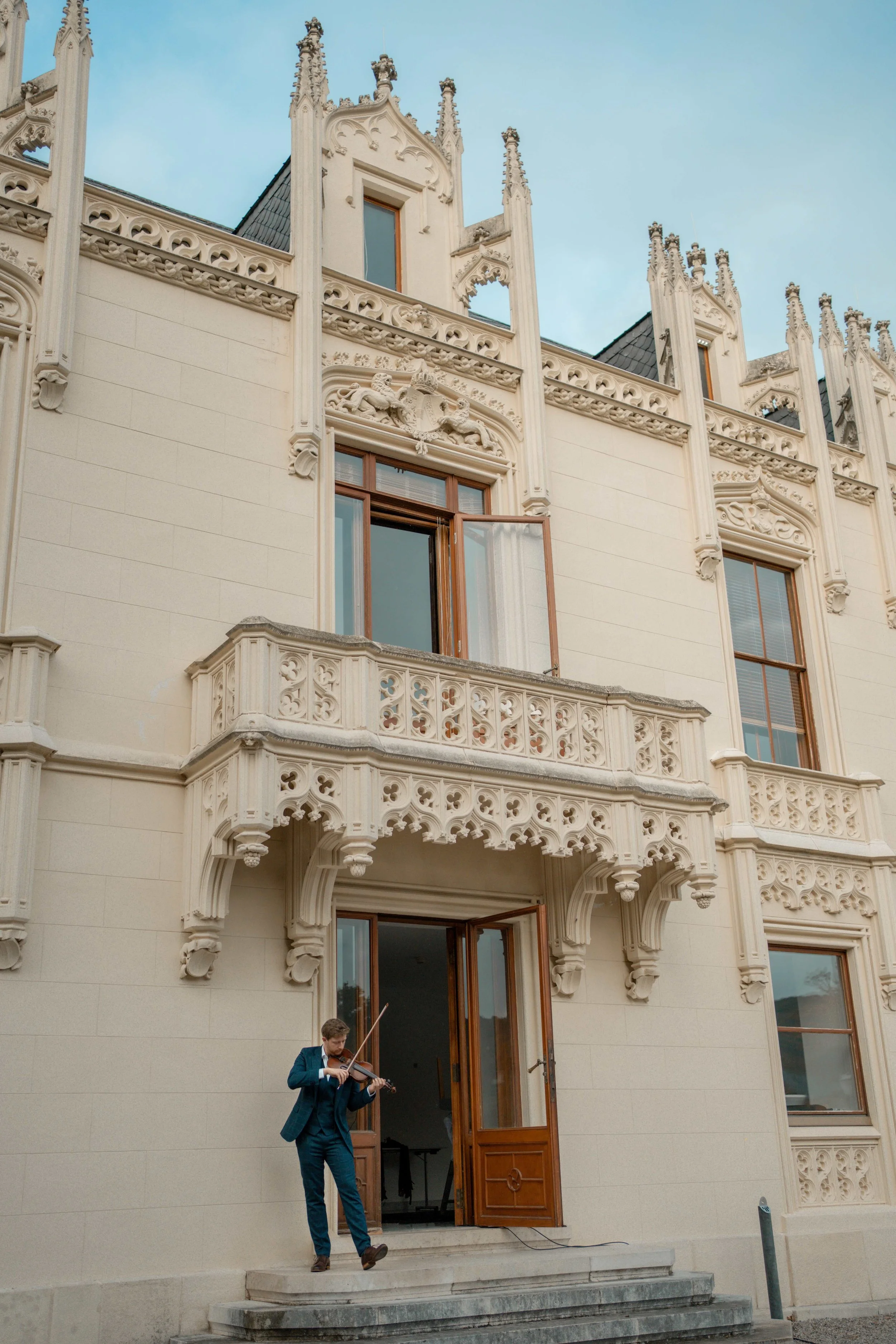 Nikolaus, in a suit playing the violin on the steps of a building with ornate classical architecture, large windows, and a balcony. The building has decorative spires and intricate stonework.