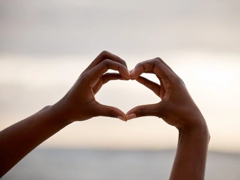 Hands forming a heart shape silhouetted against a soft sunset sky